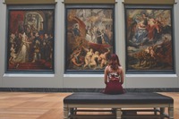 Kobieta siedzi na ławce w galerii sztuki i ogląda wiszące przed nią obrazy.
